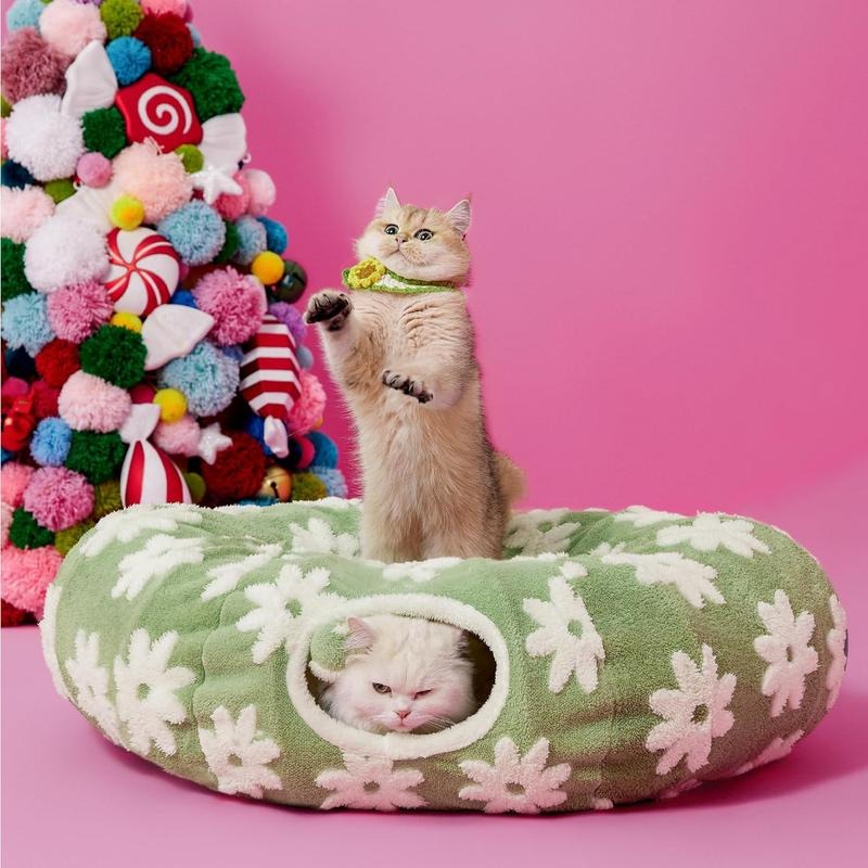 Two cats in a floral-patterned pet bed with a colorful Christmas tree in the background on a pink background